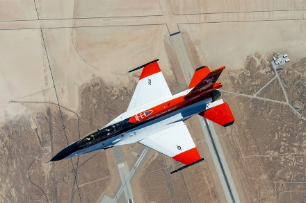 red white fighter jet flying above brown fields and air base