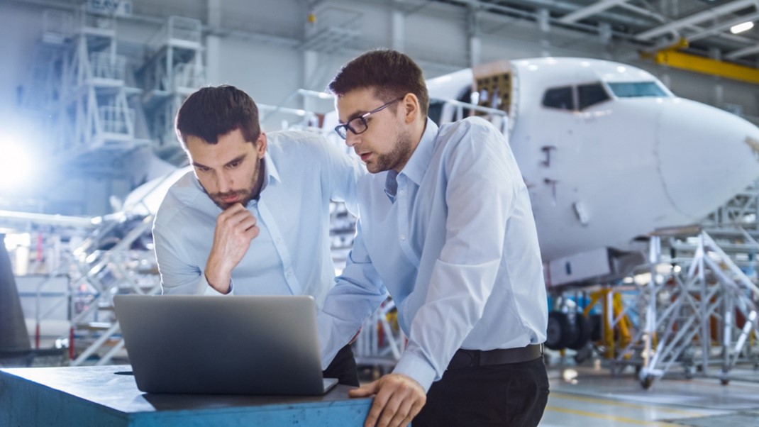 two aerospace professionals leaning over laptop in airplane hanger with airplane in background